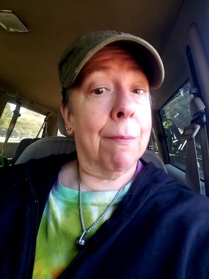 White nonbinary person in their 60s wearing a knockoff patrol-style cap, black hoodie over tiedye tshirt, amethyst crystal on silvery neck chain, and wry facial expression.