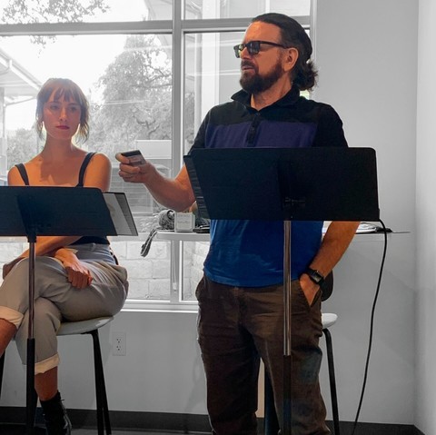 Photo of a man and a beautiful woman in front of a large window and each behind a music stand. The woman, left, is seated. The man, right, is standing, apparently reading from a card. The man has a full facial hair, sunglasses, and long hair tied into a bun.