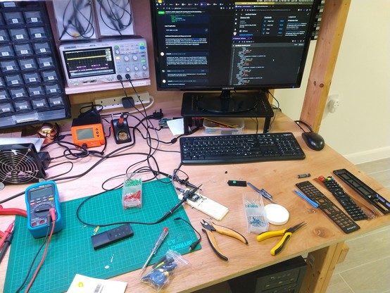 An electronics workbench containing a breadboard with esp32 stuff, multimeter, a small infrared remote for a heater, a disassembled TV remote, various small tools. There is an oscilloscope showing a yellow, mostly flat trace, and purple trace showing 2 and a half sequences of short pulses. A computer monitor shows a few windows: The largest is a github issue thread titled "remote.rc_switch supports IR receiving but not IR transmitting", further there is a window showing part of the YAML config for an esphome device, and a window with a Home Assistant configuration page on the controls the esphome device exposes (a list of buttons mirroring those on a remote).