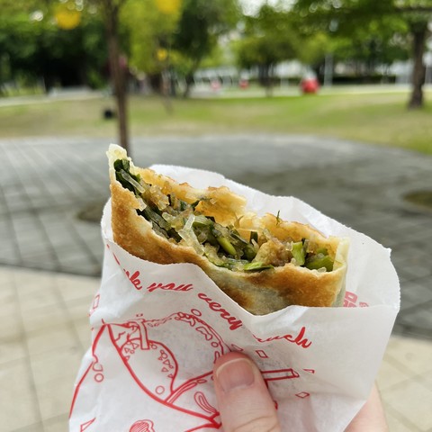 Holding a half-eaten 韭菜盒子 (“leek box”), which has a chewy doughy outer wrapping filled with green vegetables and glass noodles.  It’s being held in my hand in a white paper bag with red decorative printing on the outside and a park is visible but out of focus in the background.