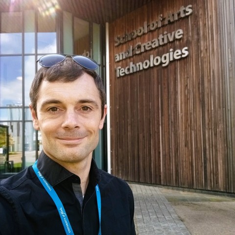 The author takes a selfie in front of the School of Arts and Creative Technologies building. Dark shirt, raised sunglasses, blue lanyard, smiling. Building consists of tall glass panels and rustic-chic faded wooden slats, with the name School of Arts and Creative technologies emblazoned in large silver letters.