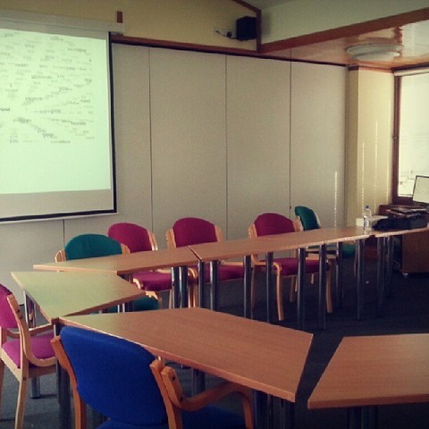 A university classroom with tables arranged into a loose circle around the room. A projector displays Dan Waber and Jason Pimble's e-poem "I, You, We", in the background.
