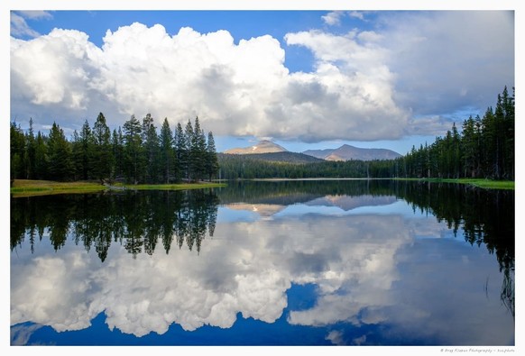 A view on a mountain lake, with dramatic white sky, fir trees and remote mountains, which reflect in the water
