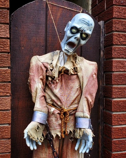 Close-up photo of a neighborhood halloween display featuring a ghoul with a blueish complexion hanging on a wooden gate. His head and torso are visible in the shot. His head leans to the right, eyes turned upwards, mouth is agape in an expression of dismay. He wears a tattered, faded red shirt with a rope tied around his waist. His arms are shackled with chains and cuffs.