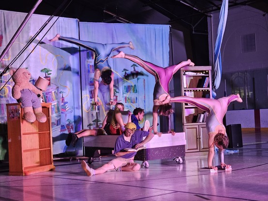 Six six performers; three as youths in colorful clothing reading while sitting on a bed prop in a stylized kids bedroom set while the other three are doing impressive handstands.