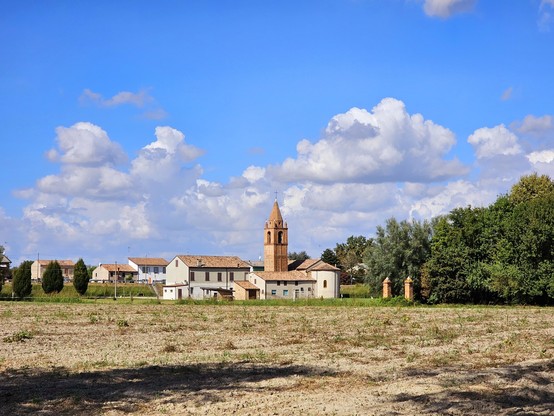 A captivating image captured in an open field, where a majestic bell tower stands in the distance. The bell tower is silhouetted against some clouds, in the sky and appears to be bathed in warm noon light. In the distance, the bell tower stands tall, with distinct architectural details sharply outlined. The surrounding field is open, with some trees on the right, and some grass. A scene that evokes tranquility and natural beauty.