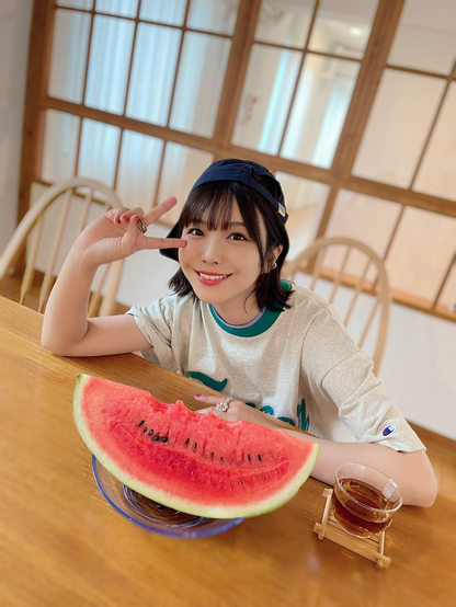 Aimi smiling doing the V sign with a watermelon on the table with some bite marks and a cup of liquid, I don’t know what it is.