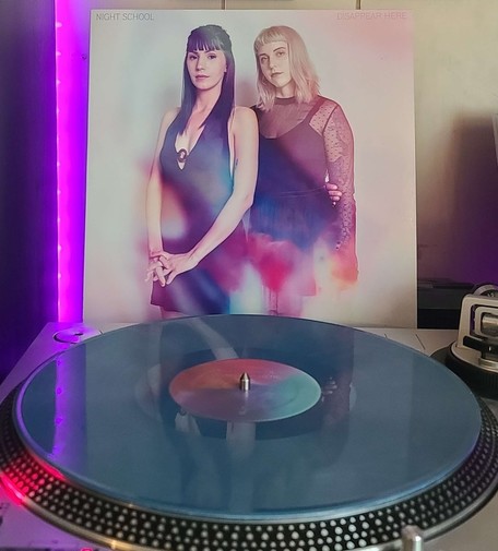 A slate blue w/ pink smoke vinyl record sits on a turntable. Behind the turntable, a vinyl album outer sleeve is displayed. The front cover shows two members of Night School standing together and looking at the camera