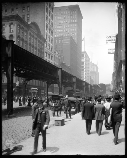 A black and white photograph along Wabash Avenue in Chicago, in 1907. Pictured is a busy sidewalk with men in suits and hats walking in both directions. A couple of horse drawn carriages are visible and an elevated train line runs down the middle of the street with buildings on either side. This photo comes from the Library of Congress.