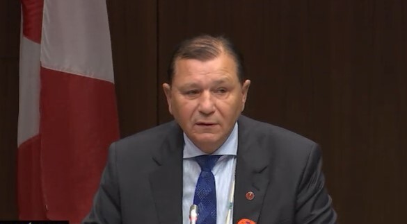 A man sits at a microphone. Behind him is a Canadian flag.