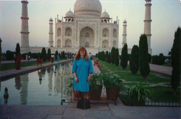 A smiling woman wearing a salwar kameez standing at the ponds in front of the Taj Mahal. The woman is somewhat out of focus and the top dome of the Taj Mahal is cut off.