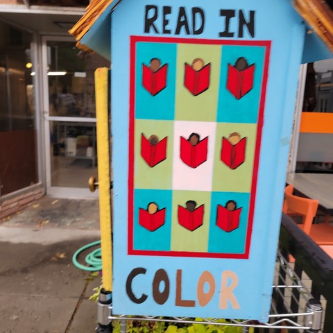 Photo of the side of a little free library painted light blue with the words READ IN COLOR and images of people with different skin colors all reading books. .