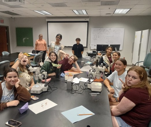 Group of happy 8th grade girls sitting around a lab bench with dissecting microscopes in front of them. One girl is pretending to drink the plankton.