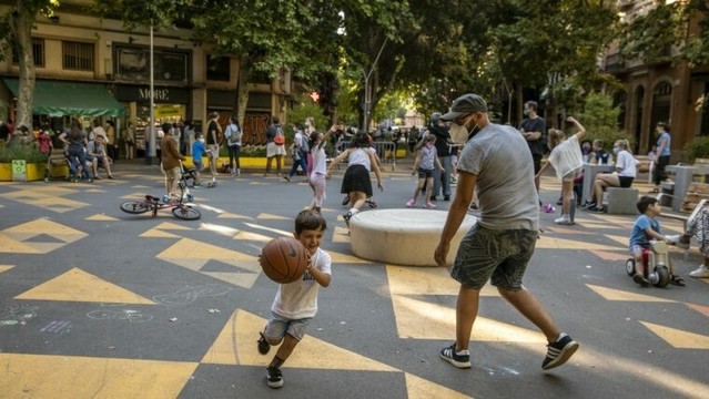 A picture of a bustling Barcelona’s street park. It’s situated on a road that’s been painted over with bright yellow markings and bordered by large planters to protect the safety of the people hanging out in the park from cars.