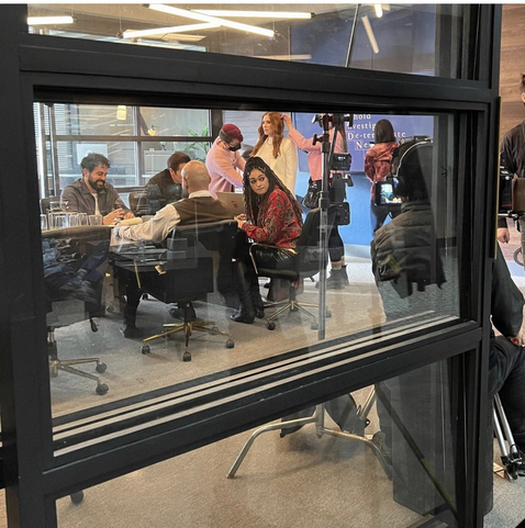 The cast of the Roman Agency are shown through a window into the boardroom used for the set.  Sarah Levy is standing for a hair and costume check while the others wait, seated around a conference table.
