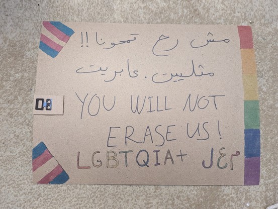 A protest sign saying:
مش رح تمحونا!!
مثلييت . عابريت
You will not erase us!
LGBTQIA+ م. ع. ل.
There's a rainbow flag drawn on the margin, and two trans flags on the corners