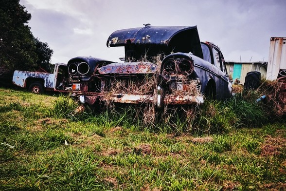 Retro car wreck, rust and grass growing through it. Bonnet arched up, headlamps empty, sky is overcast and grey.