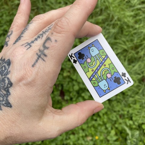 My hand, with tattoos on the back, holding a brightly coloured mini playing card between my thumb and middle finger. The king of clubs. It has a happy looking squid sort of creature on it, smiling and holding a cup of coffee.
