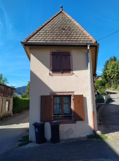 Facade of a narrow house in Buhl, France. The photo shows a wall with two small windows over each other with a gabled roof,with two dust bins in front of the house.