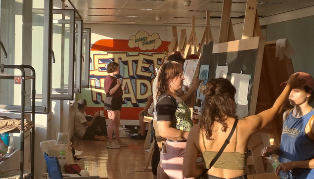 Large room with windows open to the left, a mural being painted on the far wall, and, in the foreground, people standing, talking, and painting at large a-frame style easels.
