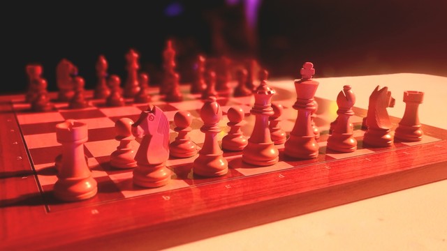 A wooden chess board with the pieces set up on a table. The pieces are throwing shadows from the stage lights