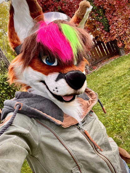 A picture of me wearing a jacket and my fursuit head standing in front of some trees with leaves that have turned a bright fall red.