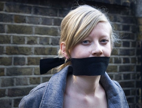 We can talk about it: A photograph of a young woman with blonde hair standing in front of a brick wall. She has a black gag tied around her mouth.