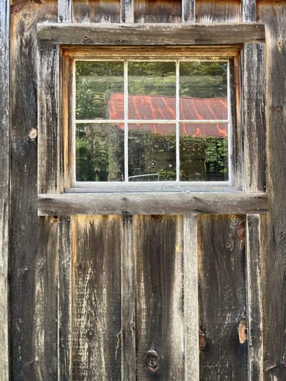 The weathered wood planks of an old barn building frame a simple six-paned window, which reflects the surrounding trees and red roof of another outbuilding opposite.