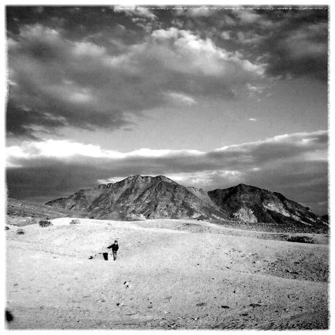 A black and white photo of a boy in a desert landscape. The boy is small and stands an afterthought in the wilderness. Skies are filled with scattered clouds.