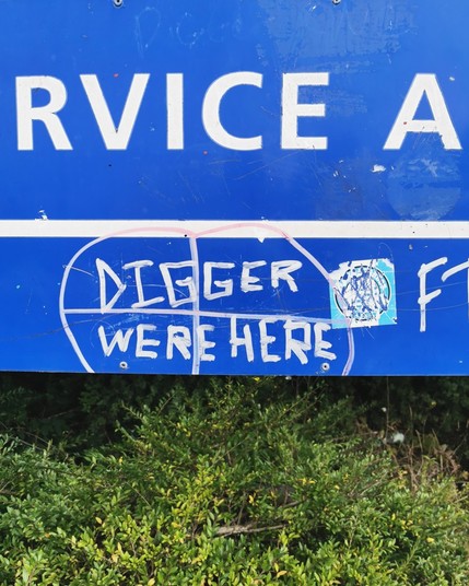 Graffiti. A blue sign for a retail park service area above some bushes. It has been graffitied with "Digger were here".