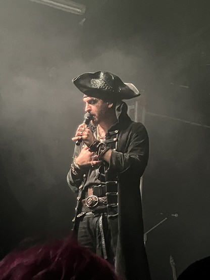 A male singer, dressed in dark pirate garb complete with tricorn hat, stands with his right hand on his chest and sings into a microphone in his left hand.