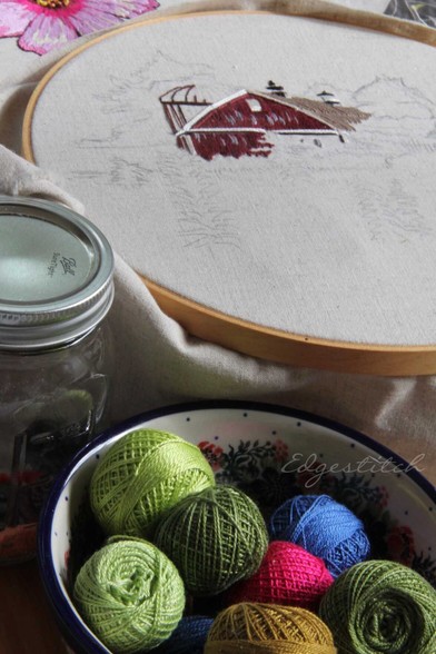 Close up "still life" type shot, embroidery hoop with partially done barn, along with some pencil sketching around it. Underneath a bowl of Valdani thread and a Ball jar with a few Dorset buttons (which you can't really see, but that's a tidbit just between us.)