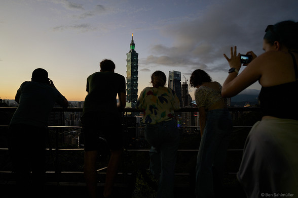 A group of tourists stands on a platform high above Taipei, taking images of the skyline at night