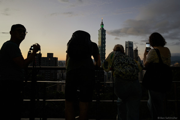 A group of tourists stands on a platform high above Taipei, taking images of the skyline at night