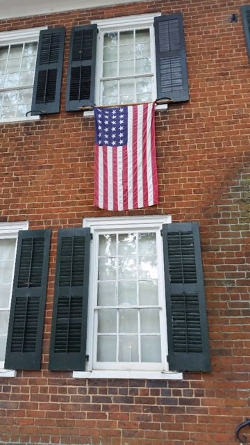 Four white painted wood windows with dark green shutters in a red brick wall. A 24 star American flag is hung vertically between the windows on the right.