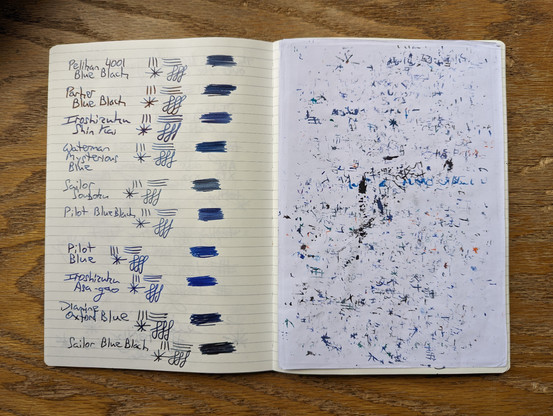 A paper notebook laid open on a wooden surface. On the right side, the page is covered by a well-used blotter sheet marked with ink. On the left side, the page lists a variety of blue and blue-black fountain pen ink names along with lines and swatches written in that ink.