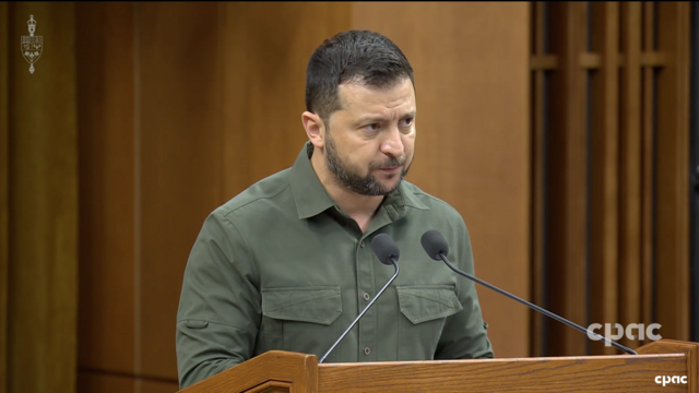 Zelenskyy in green fatigues stands behind a podium. He is looking forward with a serious face.