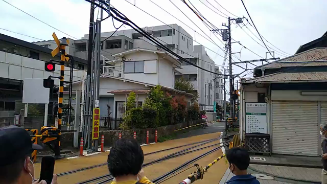 Video showing a train pulling in to Enoshima Station, Kanagawa, Japan. The video is taken to the right of the station, at the roadway crossing, which is currently blocked with safety bars, and red flashing warning lights. Additionally, a warning bell is ringing to signify the train is coming and to watch out. There are several people standing around, either waiting for the safety bars to raise or taking video of the well enjoyed Enoshima Electric Company train carriages.