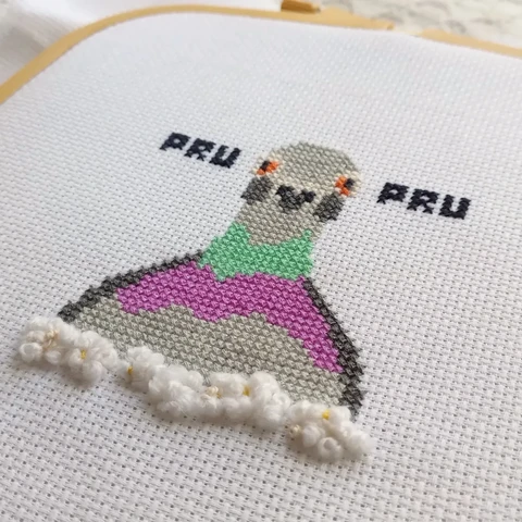 An angular photo of a cross stitch embroidery of a pigeon and some pop corn made in french knots with wool.