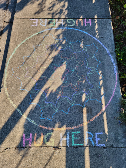 A chalk art pattern of our latest hug zone, created using two diamonds in a revolving pattern. At the edges of the drawing are the words HUG HERE