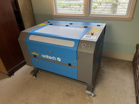 An industrial size laser cutter sitting in my house. It is bright blue and grey and says Omtech on the front in white letters.