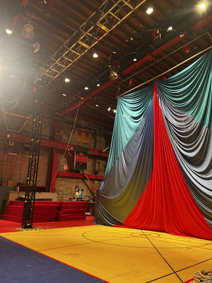 A yellow floor with lines criss-crossing it geometrically. At rear, a large teal, blue and red curtain draped with gentle waves through the fabric. Brick wall, warehouse. A silver truss near the roof. A sense of spaciousness. A sign on the back wall reads, “España-Streb Trapeze Academy.”