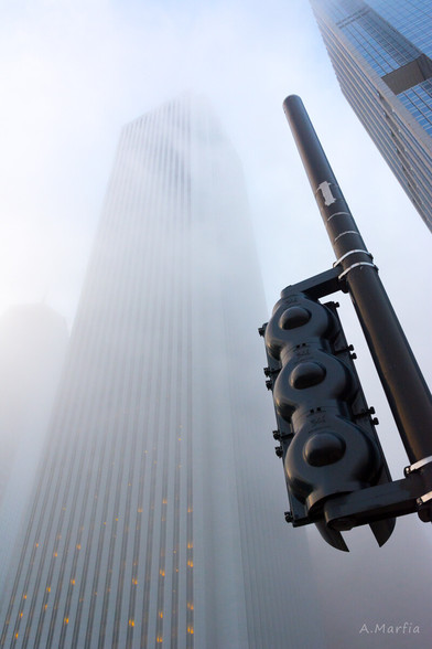 A color photograph in Chicago on a foggy day. Pictured is a street light juxtaposed with the Aon Center skyscraper. Some lights can be seen in the lower level windows while the upper floors are enveloped in fog. Circa 2014.