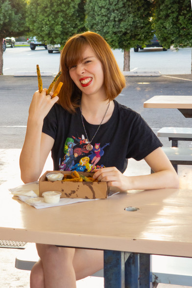 Photograph of Lety seated in the outdoor dining section of a burger place with a paper tray of fresh fried zucchini on the table in front of her. She's wearing a black Bruce Timm "Women of Marvel" t-shirt and denim booty shorts. Lety has three fried zucchini in between the fingers of her right hand. She grits her teeth, partially to commit to looking like the Wolverine and partially because she feels hot oil dripping down her palm.