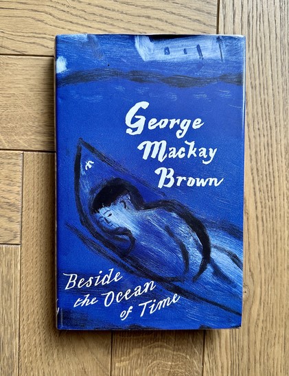 George Mackay Brownâ€˜s 1994 novel Beside the Ocean of Time lying on a wooden floor. The cover shows an illustration by Charles Shearer, in blue, white and black, of a stylised figure in a small boat or coracle with a whitewashed cottage just visible on the shore in the background. The novel was Brownâ€™s last before his death in 1996, and was shortlisted for the Booker Prize and awarded Scottish Book of the Year by the Saltire Society.