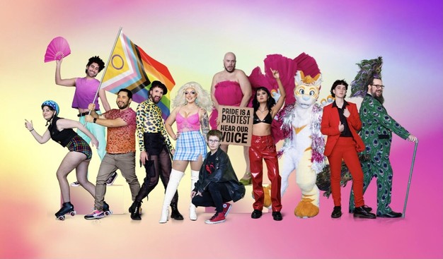 This image shows a group of eleven people, all wearing different  clothing and footwear expressing their own individual LBGTQIA+ pride outfit. The majority of the people are wearing bright colors such as pink, magenta, and red. There is one person on the left who is holding up an inclusive rainbow flag. To their right is another person with long black hair wearing a black jacket and red pants holding a sign reading “Pride is a protest. Hear our voice.”  To the right of this is an individual in a full body Furry costume. At the far right there is the author  wearing a three piece suit with a peacock pattern on it, a peacock Mohawk and a flowing peacock tail. They are posing using their point stick to lean on. Everyone posing for the photo are, smiling at each other or looking directly into the camera lens with joyous expressions on their faces.