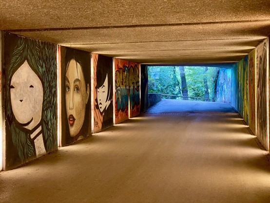 Long and wide cement tunnel. At the end we see blue-green light, and to the left is a series of artists paintings