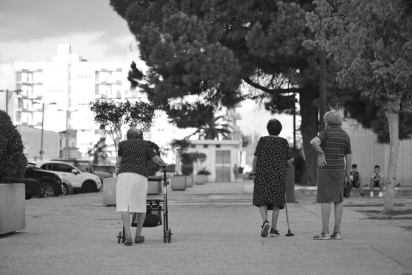 drei ältere, weiblich gelesene personen laufen durch eine spanische stadt über den gehweg. sie sind von hinten fotografiert.
die linke person mit rollator, die mittlere mit gehstock und die linke person trägt handtasche und hat eine hand in die seite/rücken gestützt.
im hintergrunf sitzen jugendliche auf einem treppenabsatz
