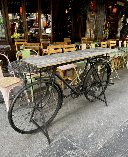 Cafe sidewalk table made from a bicycle topped with wooden boards. Cafe tables and chairs surround it, restaurant windows on background.