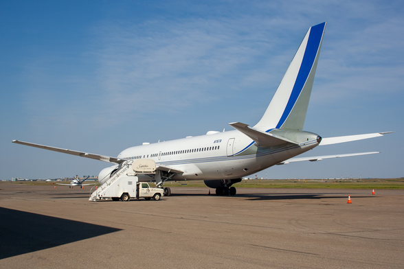 A twin engine jet liner parked facing away to the left, painted white with a blue and gray swish up the tail. There is a stair truck parked to the left of the plane. The sky is blue with wispy clouds. Orange pylons are to the right of the plane.
